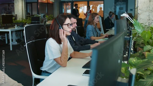 Female wearing headset sitting in busy office workspace. Listening to client while typing on keyboard. Colleagues focused on computers nearby. Support team handling communication in dim office.