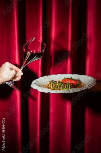 A hand holds a unique glass of red wine with a creative angle next to an elegant plate of gourmet food, all set against a lush, dramatic red velvet curtain.