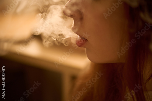 Caucasian young adult woman exhaling vapor from mouth, closeup showing side of face and lips with visible smoke, indoor setting, focusing on vaping action and facial features