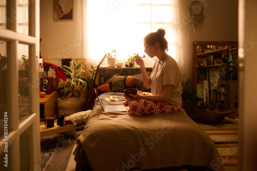Caucasian young adult woman sitting cross legged on bed eating breakfast while working on laptop in cozy bedroom, with plants and personal items visible in background