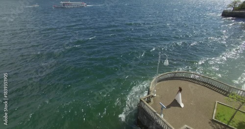 A high-angle aerial drone shot of a bride in a flowing white wedding dress gracefully walking towards a wooden pier on a beautiful lake shore. Majestic mountains form a stunning, cinematic backdrop. 