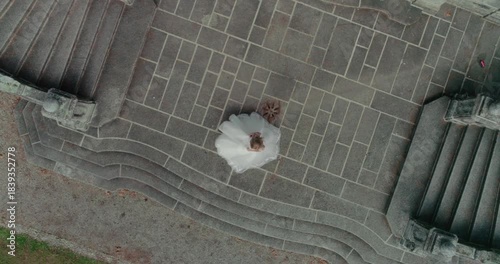 A high-angle drone shot looking down at a bride in a white wedding dress joyfully spinning on ancient, textured stone stairs. Captures movement, romance, architecture