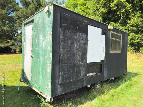 Tableau sur toile An old locked wooden storage shed or cabin or hut in poor condition, with peeling paint, standing on a trailer, in a grass field