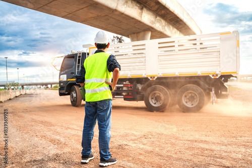 Bild auf Leinwand Construction Worker Overseeing Truck on Construction Site Near Highway Under Dra
