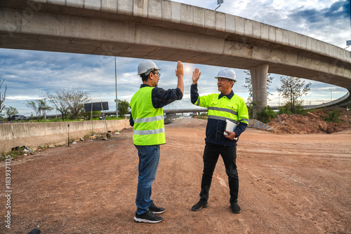 Two Construction Workers High-Fiving Under a Bridge on a Construction Site During Sunset