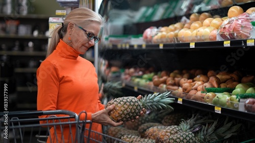 A mature woman examines a pineapple in a grocery store while comparing fruits and considering her budget for healthy meals.