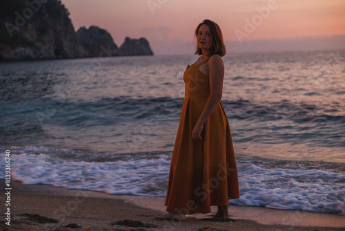 Woman On Sandy Beach At Sunset, Standing Barefoot In Flowing Rust Dress, Waves Lapping, Dramatic Cliffs
