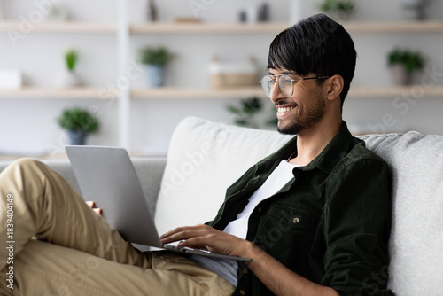 Photos A young man sits comfortably on a couch with a laptop on his lap, looking engaged and smiling