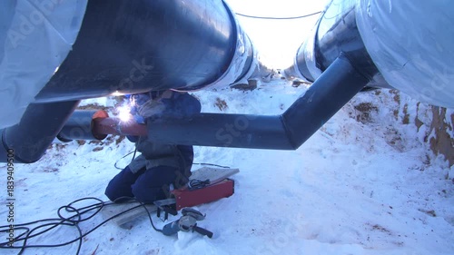 Welder working on heating pipeline construction in winter outdoors.