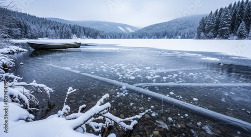 Solitary rowboat rests near the partially frozen shore of a serene winter lake surrounded by snow-covered slopes.
