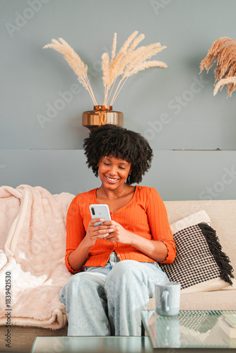 Joyful woman engaged in using her smartphone while sitting comfortably on the couch at home, smiling and feeling content in her personal space, showcasing a moment of leisure and connection.