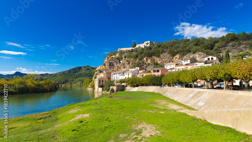 Scenic view of Miravet village and castle by the Ebro river in Catalonia, Spain, with green grass and blue sky.