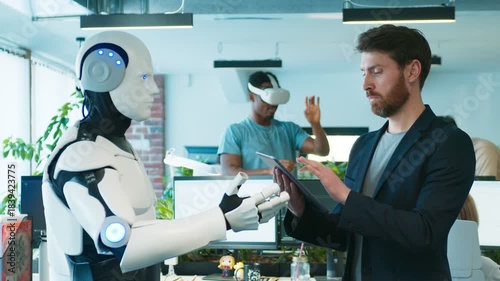 Male testing humanoid robot. Developer using tablet while robot extending hand. Coworker in background putting on VR headset. Busy workers sitting at desks and looking at monitors.