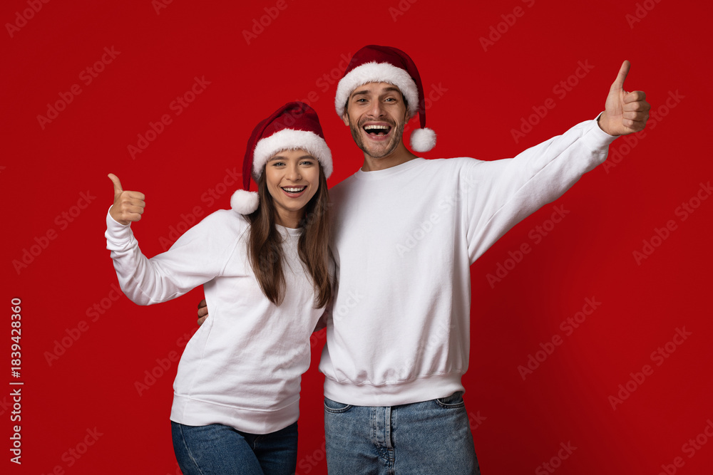 Fototapeta premium A happy couple wears matching Christmas hats while posing against a bright red background. They express joy with smiles and thumbs up, embodying holiday spirit.