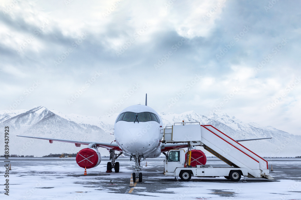 Obraz premium White passenger plane with boarding stairs on the winter airport on the background of high snow capped mountains