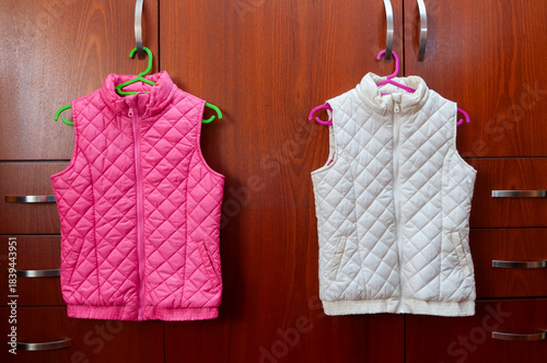 Two quilted vests, one pink and one white, hanging on hangers above a dark wood wardrobe.