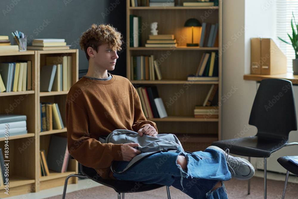 Fototapeta premium Teenage boy sitting alone in group therapy room holding backpack on lap looking to side appearing thoughtful surrounded by bookshelves and empty chairs