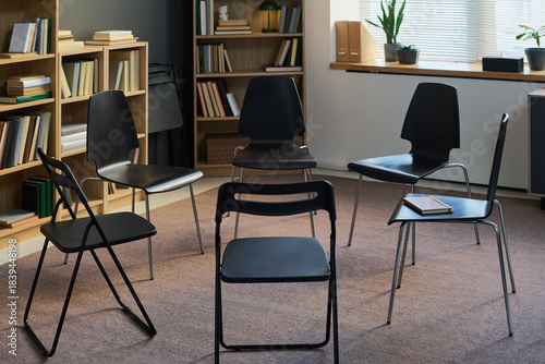 Empty chairs arranged in circle in therapy room suggesting group therapy session setting, books placed on seats and shelves indicating preparation for discussion or support meeting
