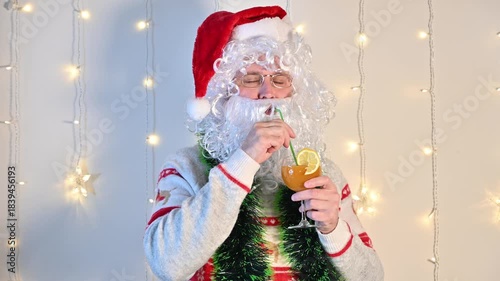 A man dressed as Santa Claus on a white background with festive lights, enjoying a tropical cocktail with a citrus slice and a drinking straw, creating a fun and relaxed holiday scene.