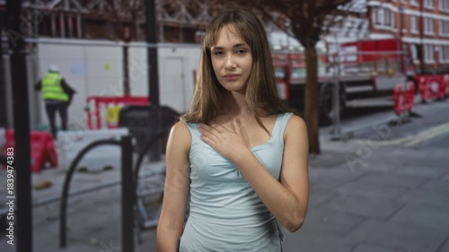 Young woman with hand on chest on street at a construction site, wearing light blue sleeveless top and neutral expression; quiet confidence.