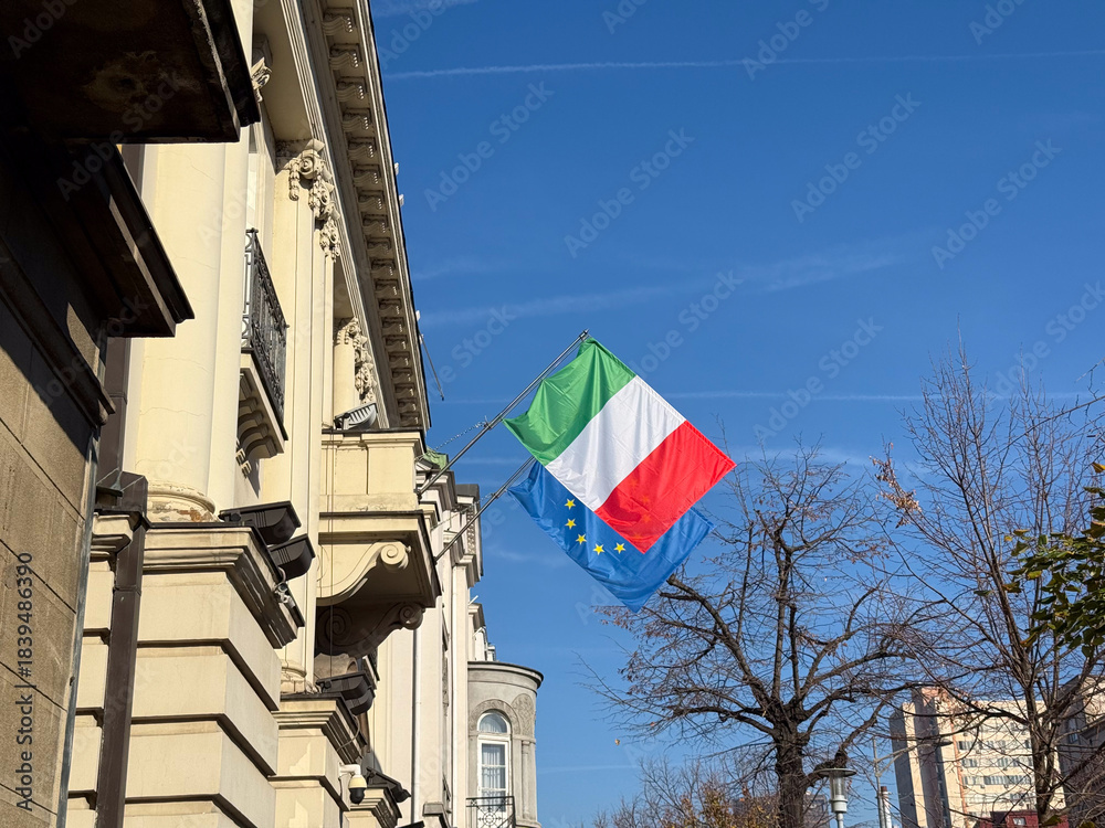 Naklejka premium Italian and European flags waving near historic building under clear sky. Symbol of cultural connection, diplomacy, political unity and national identity in urban environment.