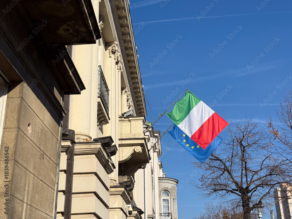 Naklejka premium Italian and European flags waving near historic building under clear sky. Symbol of cultural connection, diplomacy, political unity and national identity in urban environment.