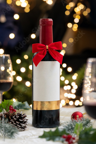 A bottle of red wine and red ribbon bow with Christmas decoration, light illumination, festive ambiance, glass of wine, illuminated light bokeh in background.
