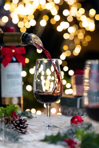 A clear glass of red wine while bottle pouring into glass, festive ambiance and Christmas decoration atmosphere with bokeh light illumination in background.
