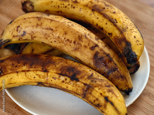A bunch of overripe bananas with brown spots resting on a white plate on a wooden table. Close up.