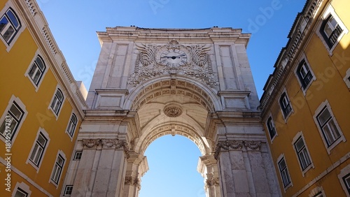 Elaborate stone archway under blue sky, flanked by yellow buildings, centered