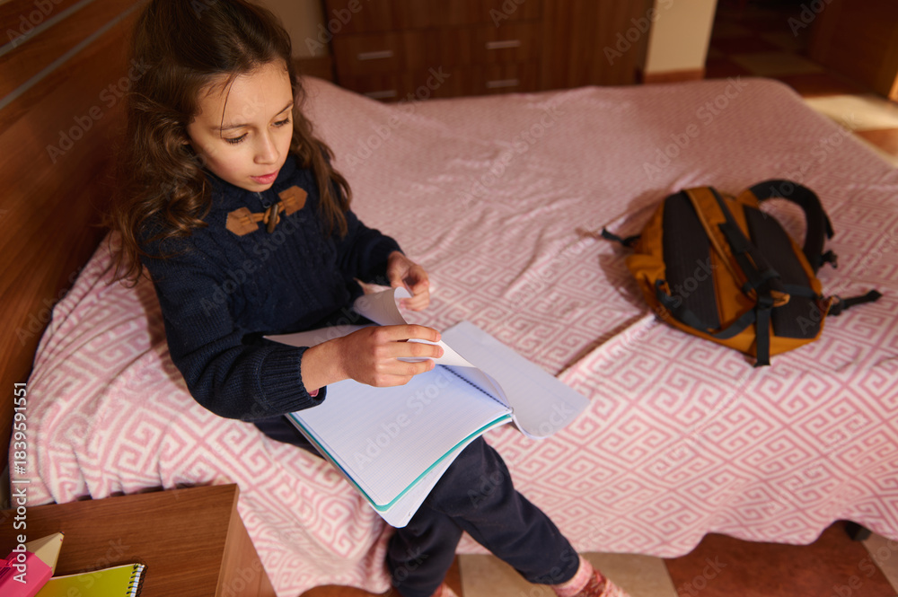 Obraz premium View from above of an elementary age student girl engaged in homework at home, sitting on her bed. A backpack is nearby