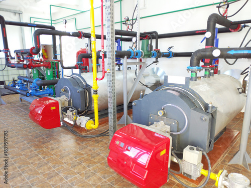 A modern boiler room with two large industrial boilers. The boilers are connected to pipes supplying water and gas. The pipes are equipped with pressure gauges, shut-off valves, pumps, and fittings.
