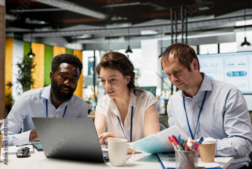 Fotografie Multiethnic corporate team, including black man and caucasian individuals, collaborating in office, using laptop and documents to assess investment reports and business analytics