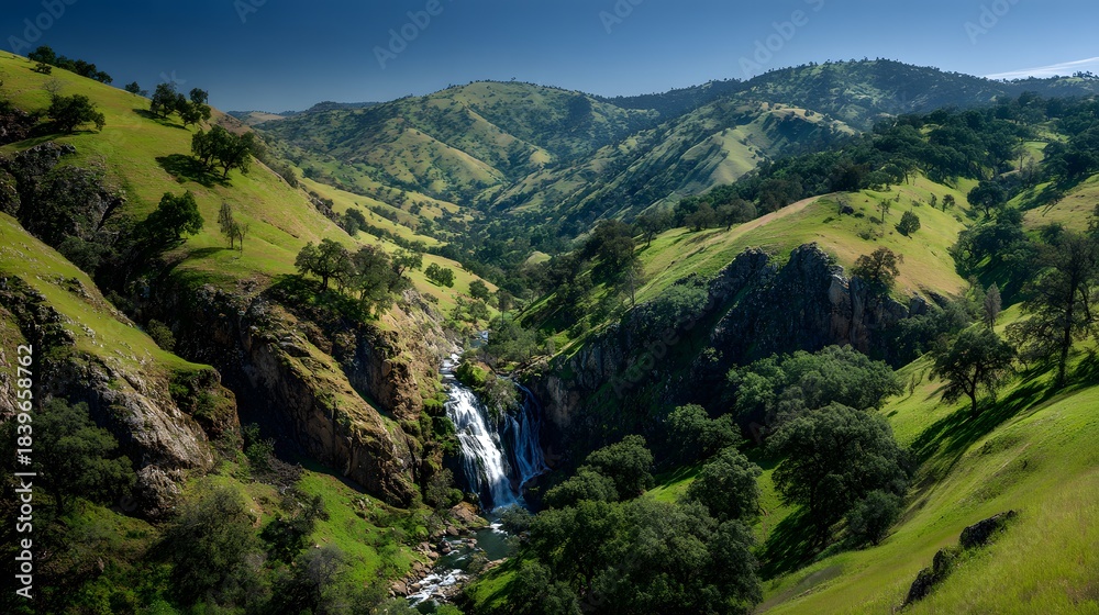 Fototapeta premium Lush green rolling hills cradle a cascading waterfall flowing through a deep ravine under a clear blue sky