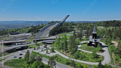 Holmenkollen Ski Stadium 4K Drone Top Angle, Oslo, Norway. Tourists walking arena paths below the ski jump tower, open sunny day, geometric stadium layout.
