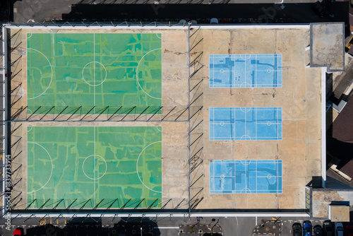 Aerial top-down view of a rooftop sports facility with two large green futsal courts and three smaller blue sepak takraw ball courts, enclosed by fencing and surrounded by an urban residential area.