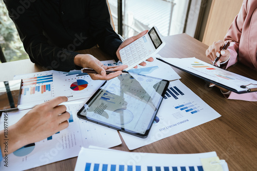 A group of professionals reviews charts on paper and a tablet, discussing financial data and calculating figures during a business meeting.