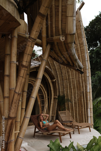 A woman rests on a lounge chair outside a bamboo eco villa overlooking lush tropical greenery and handcrafted wooden architecture