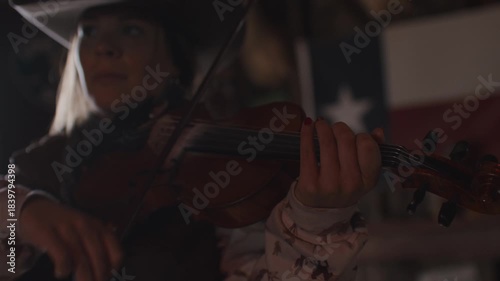 Close Up of Woman Wearing Cowboy Hat Playing Fiddle Violin Music In Front of Texas Flag Inside of Old Rustic Shadowy Saloon