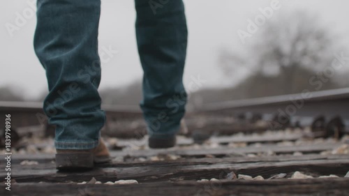 Man Wearing Jeans and Cowboy Boots Walking Along Old Rural Railroad Tracks With Overcast Misty Foggy Weather