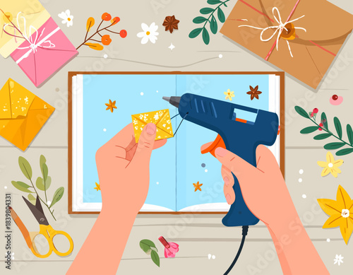 From a top-down perspective, hands use a hot glue gun to attach a small envelope to a scrapbook. The workspace is creatively cluttered with scissors, paper, and floral elements, perfect for a DIY.