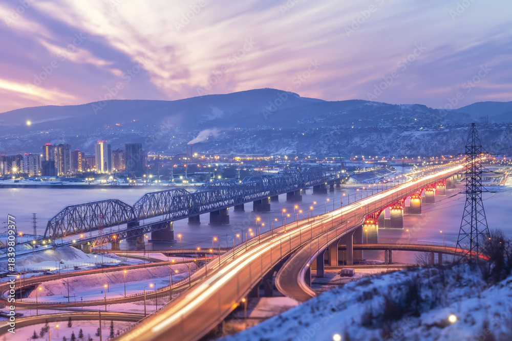 Fototapeta premium Beautiful bridges in the night illumination. Nikolaevsky Fourth Bridge in Krasnoyarsk, Russia and the railway bridge over Yenisei River. Traffic and urban transport movement, Siberian city in winter
