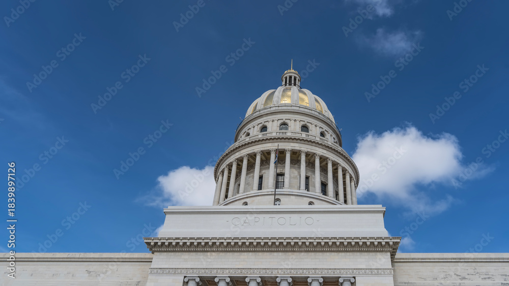Obraz premium The Capitol Building, Havana. Cylindrical tower with colonnade, carved capitals, balustrades, arched windows, gilded dome and spire against a blue sky, clouds. The inscription on the facade : Capitol
