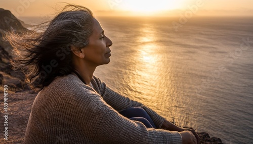A woman sits on a cliff overlooking the ocean at sunset, her face turned towards the warm golden light.