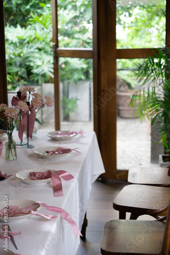 An elegantly set table adorned with pink ribbons and delicate flowers, bathed in natural light from large windows overlooking lush greenery