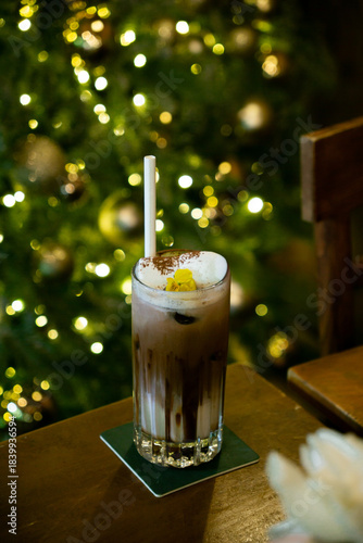 A festive hot cacao topped with marshmallow and a yellow flower garnish, set against a sparkling Christmas tree backdrop.