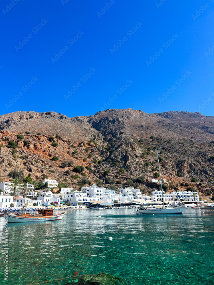 Obraz premium Loutro coastal village in Crete island Greece. White houses, moored boats on calm water below rocky hills, blue summer sky
