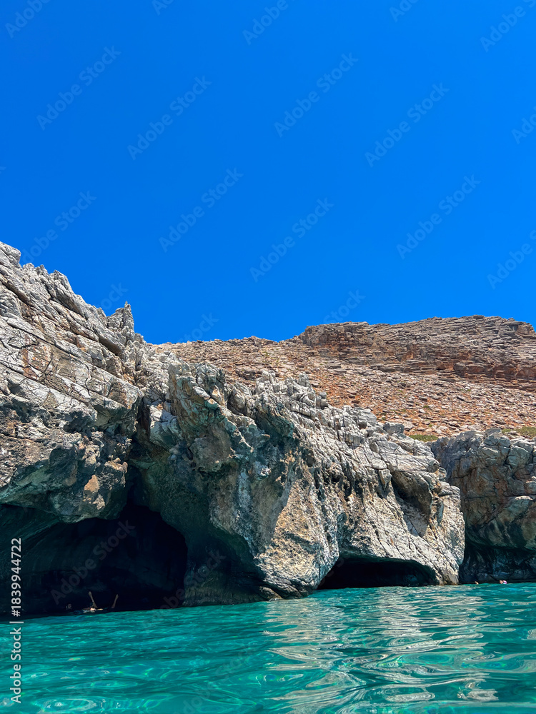 Naklejka premium Sea cave with turquoise crystal water along the rocky Crete coast, Greece. Blue summer sky, bright reflections on water