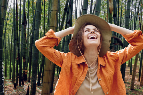 Woman wearing a hat laughing in a bamboo forest, outdoor nature portrait with joyful smile, casual orange jacket and relaxed carefree travel vibe captured in sunlight.