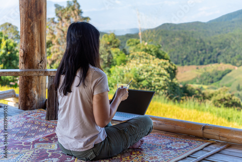 A female digital nomad working on her laptop and enjoying a coffee break while sitting on a mat with a scenic mountain view in rural Asia. Conceptual image for remote work and work-life balance.
